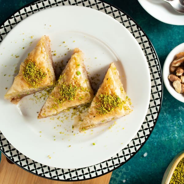 Three triangular baklava pieces, topped with crushed pistachios, on a white plate with a patterned rim.